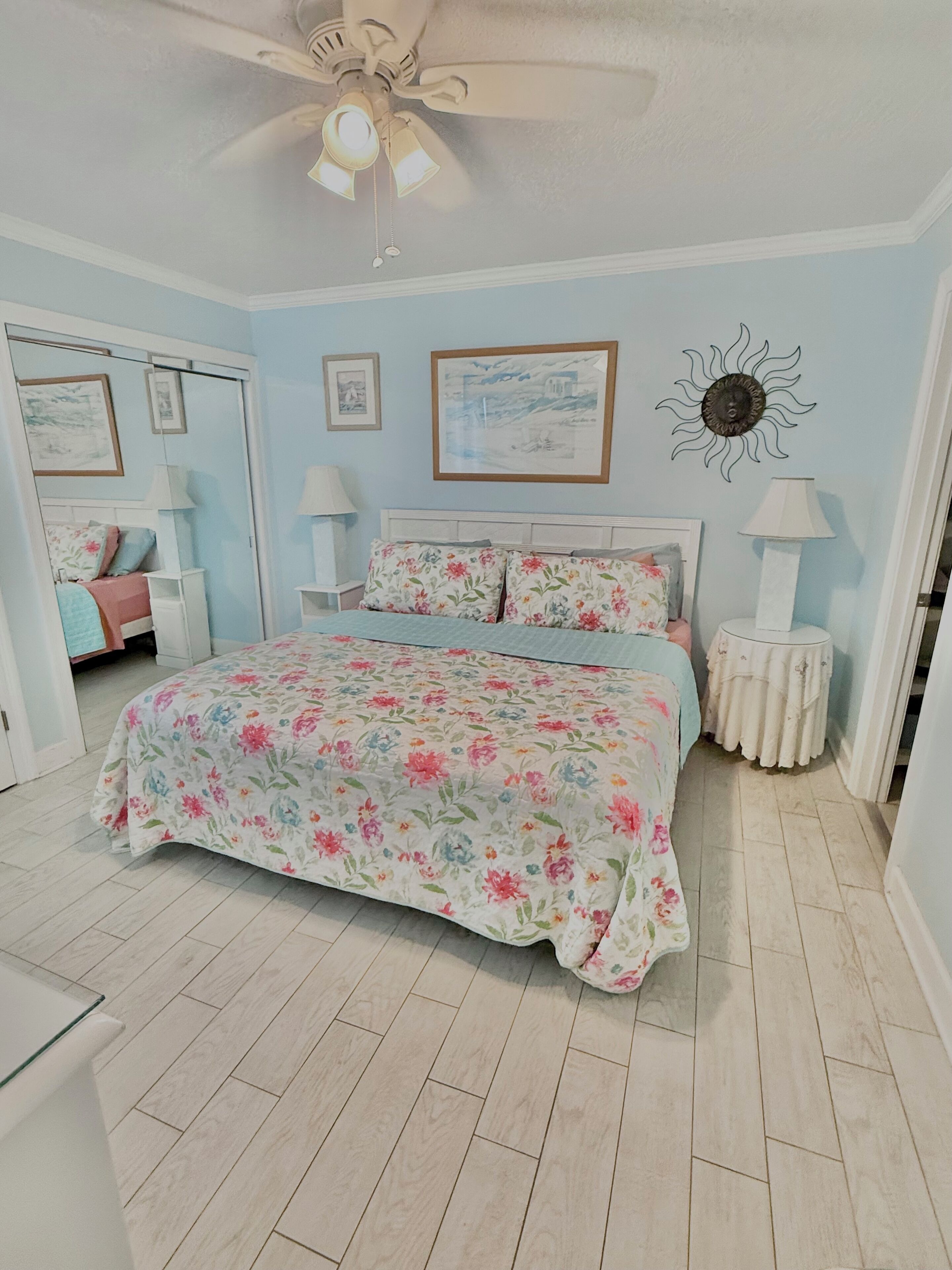 A bedroom with a floral bedspread, a ceiling fan, and a sun-shaped wall decoration.