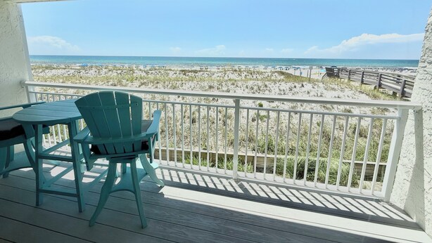 A balcony with two chairs and a table, overlooking a beach with a wooden fence and beachgoers.