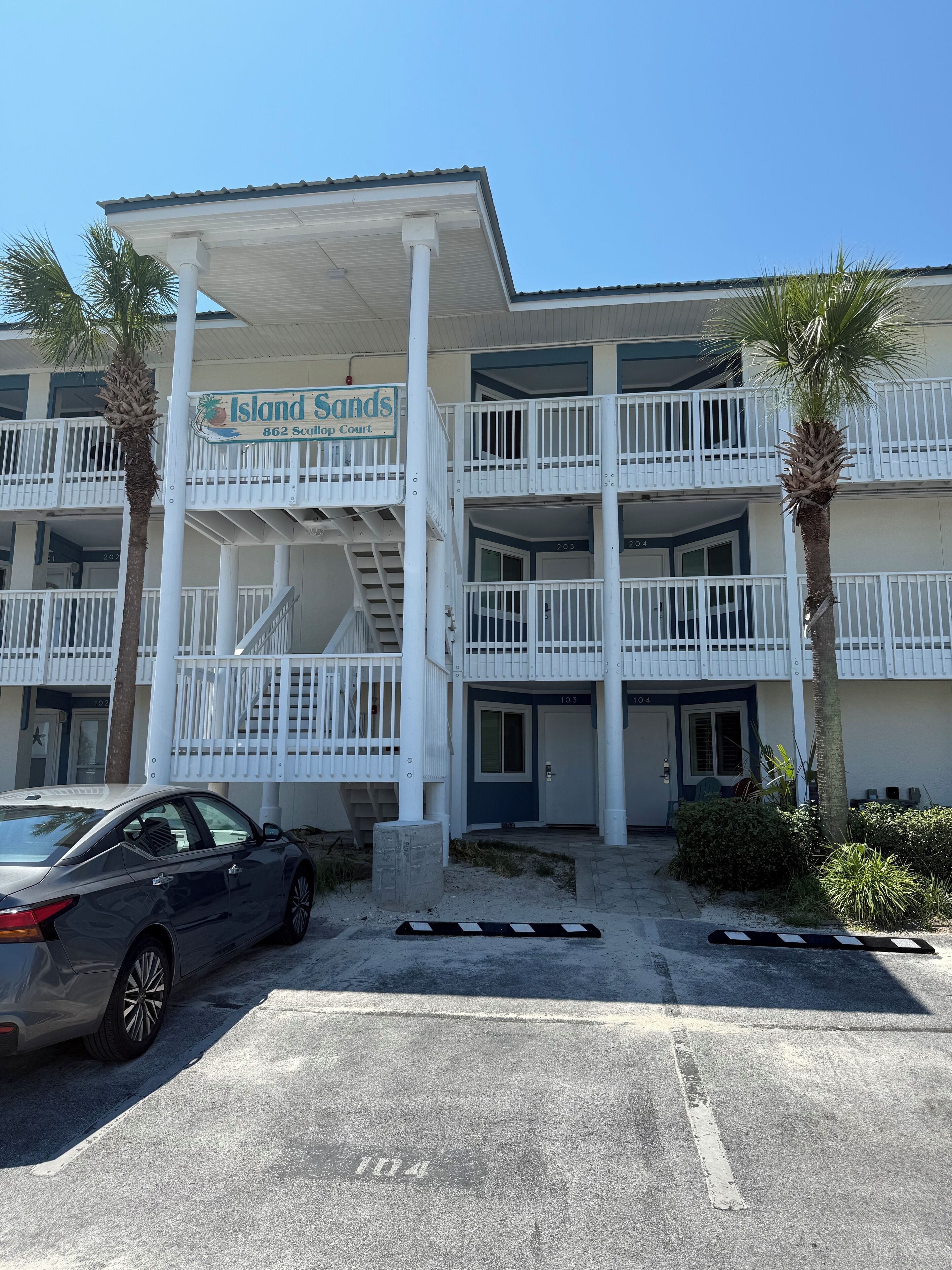 A multi-story hotel with balconies, a car parked in front, and palm trees.