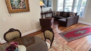 Un coin repas avec une table ronde, des chaises et un vase de fleurs. Le salon comprend un canapé et un téléviseur, et il est doté de grandes fenêtres.