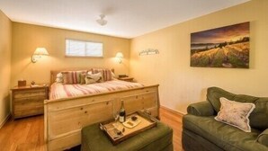 A bedroom with a wooden bed, bedside lamps, a green armchair, a painting on the wall, and a window with blinds. - Comfort and Style in Our Queen Cottage (Calistoga)