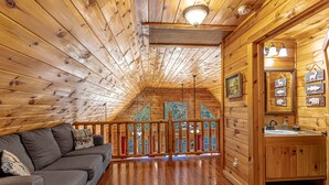 Interior de una cabaña de madera con techo de madera, un sofá, ventana con marco de madera y un baño de madera con lavamanos y espejo.