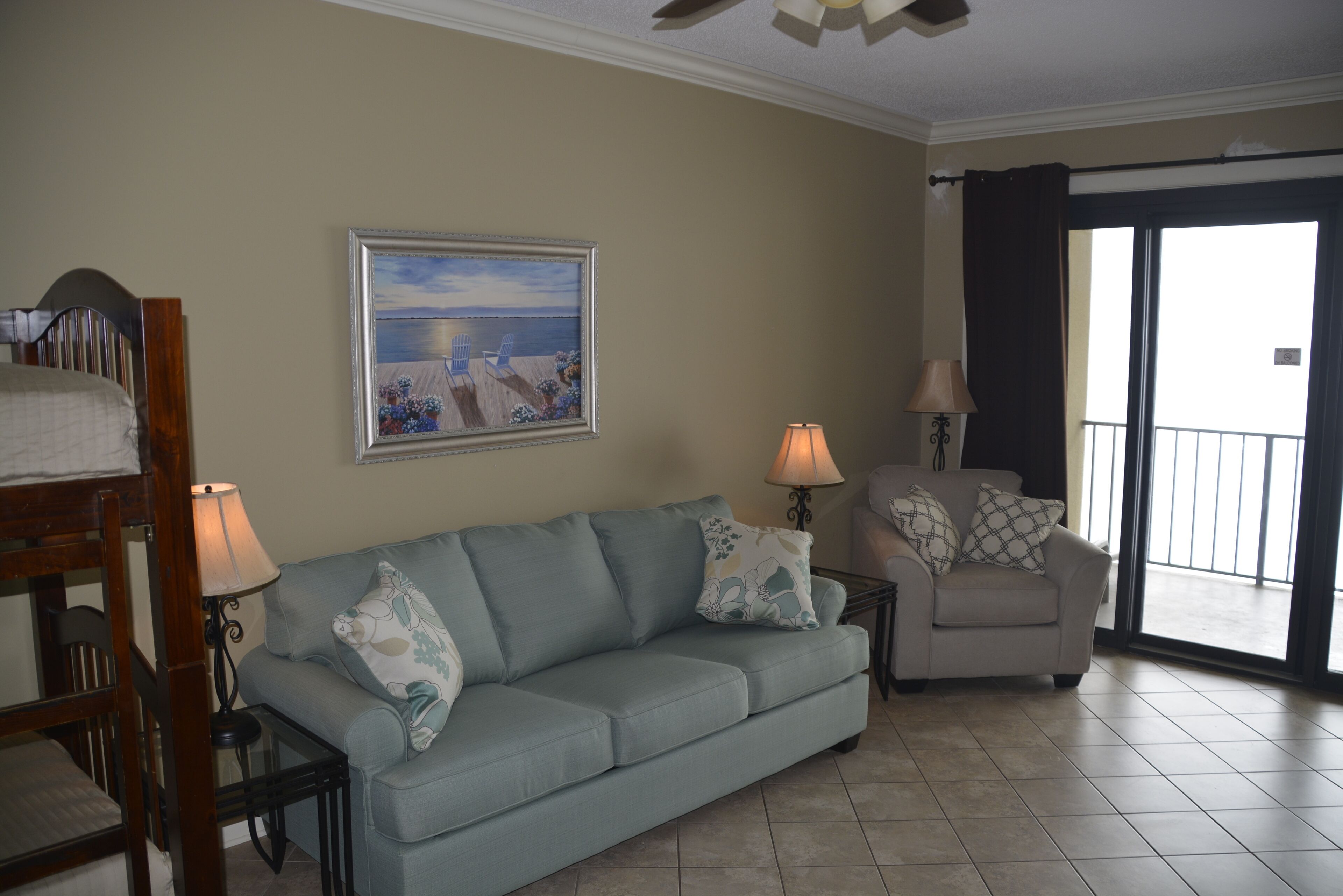 A living room with a light blue sofa, two side tables with lamps, a framed painting, and a balcony with a view.