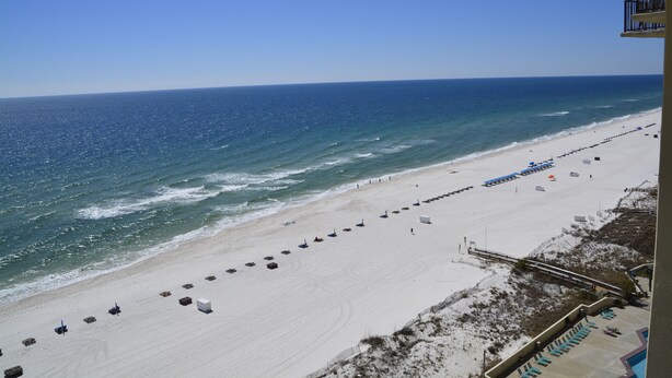 Ein Strand mit einer langen Reihe aus Sonnenschirmen und Liegestühlen, freier Blick auf das Meer und ein Gebäude mit Balkonen mit Blick auf den Strand.
