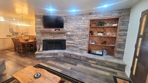 A living room with a stone fireplace, a flat-screen TV mounted above it, and a wooden shelf with decorative items.