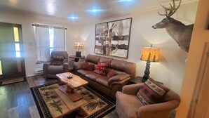 A living room with a brown leather sofa, two armchairs, a coffee table, and a rug with forest patterns. There is a deer head mounted on the wall, a lamp with a tree design, and a painting above the sofa.