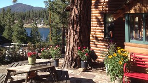 A wooden cabin with a stone patio, surrounded by pine trees and a view of a lake and mountains. - First-Tier Lakefront Log Charmer! Lakeview Spa In Gazebo,Fireplace,Deck,Sledding (Big Bear Lake)
