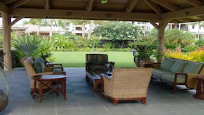 A covered outdoor seating area with wicker furniture, a coffee table, and a view of a well-maintained lawn and buildings in the background.