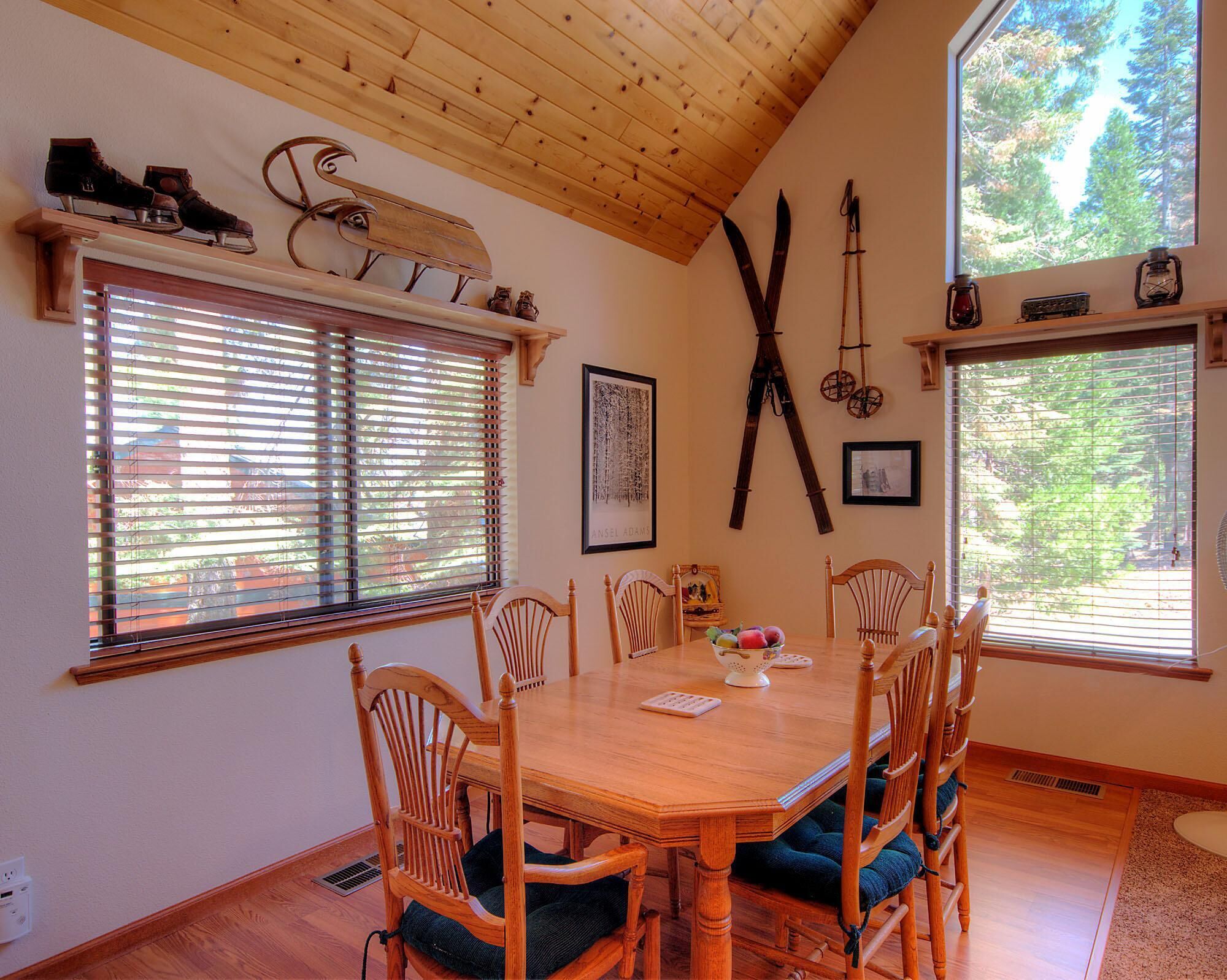 Un coin repas avec une table et des chaises en bois, des poutres apparentes au plafond et de grandes fenêtres équipées de stores.