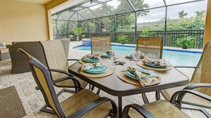 Área de refeições ao ar livre com mesa posta para quatro pessoas, com vista para uma piscina e vegetação exuberante.