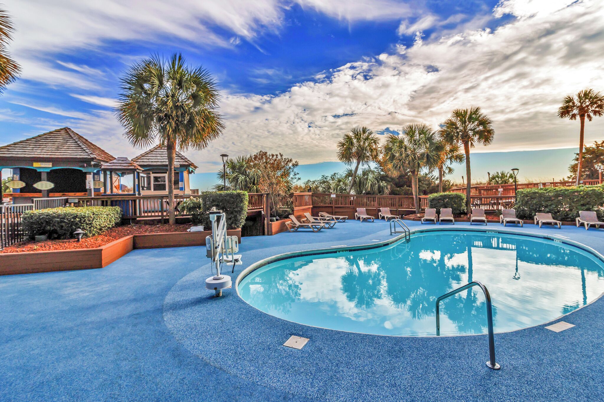 A pool area with lounge chairs, palm trees, and a building with a roofed deck.