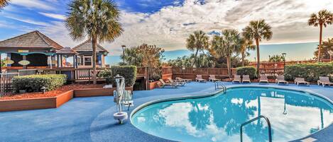 A pool area with lounge chairs, palm trees, and a building with a roofed deck.