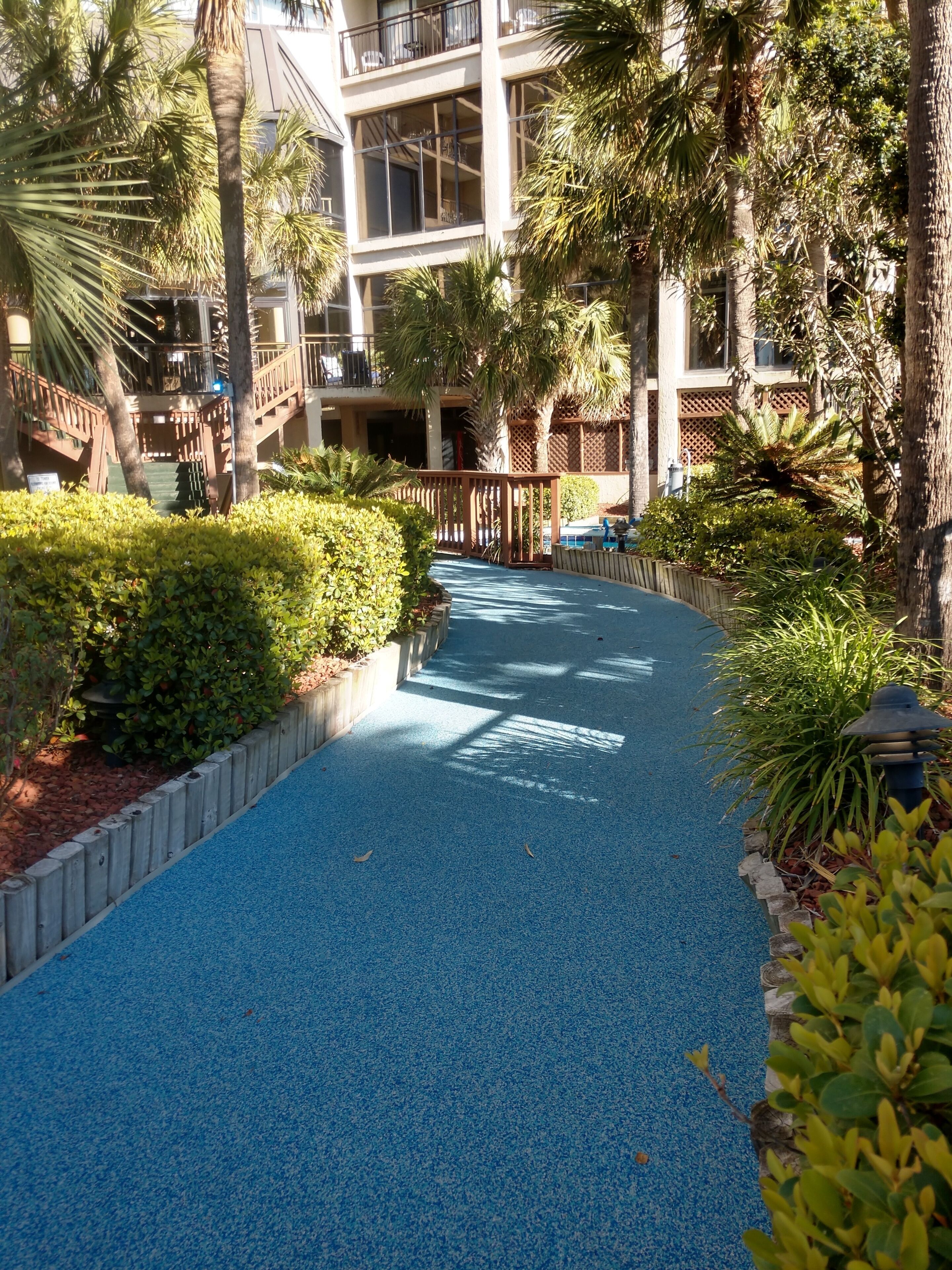 A pathway with blue tiles, surrounded by greenery and a building with balconies.