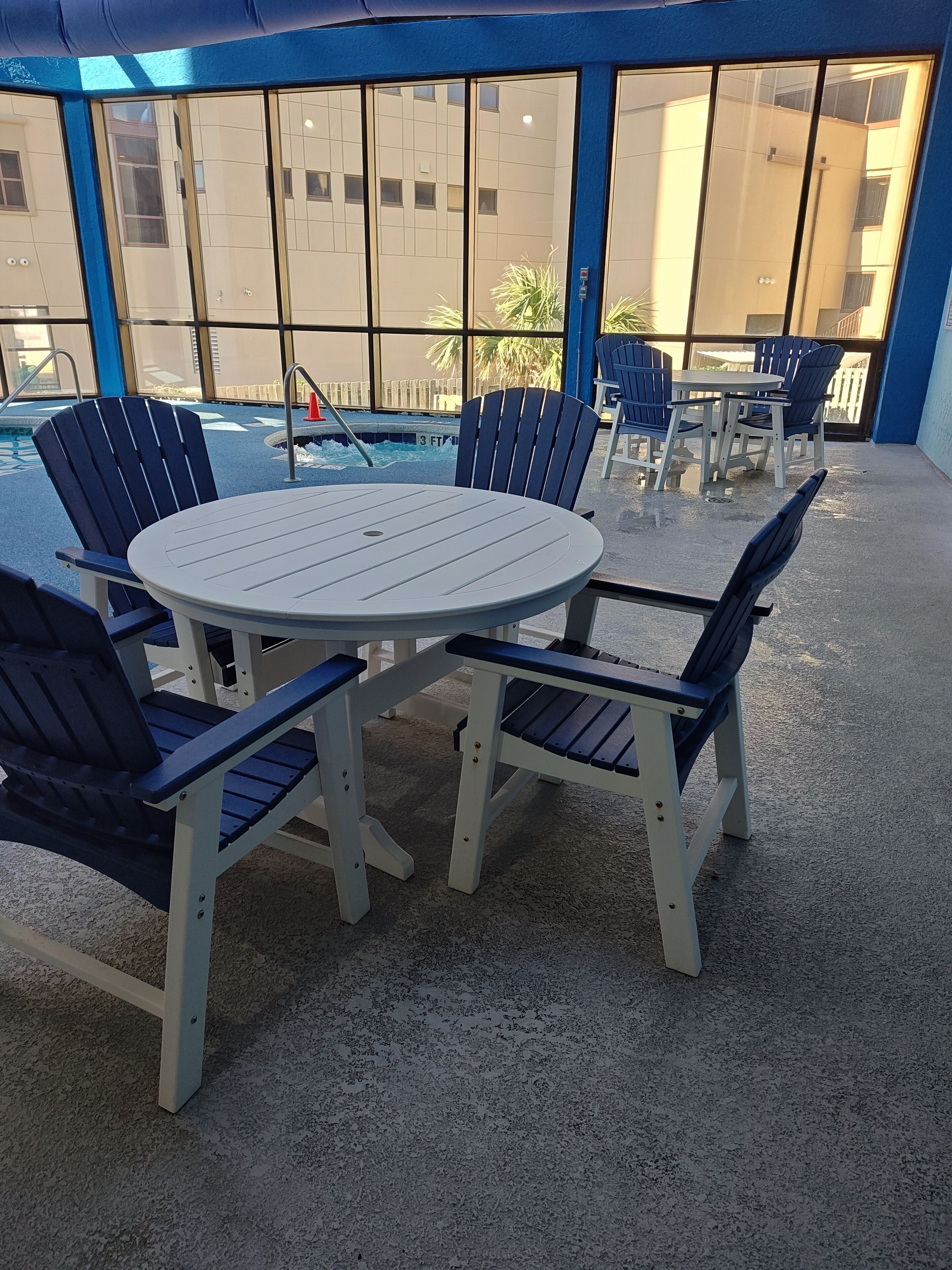 A covered outdoor seating area with a round table and chairs, a pool in the background, and large windows.