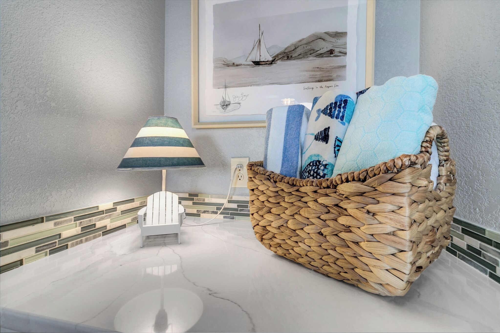 A wicker basket with towels on a marble countertop, a lamp, and a framed picture of a sailboat on the wall.