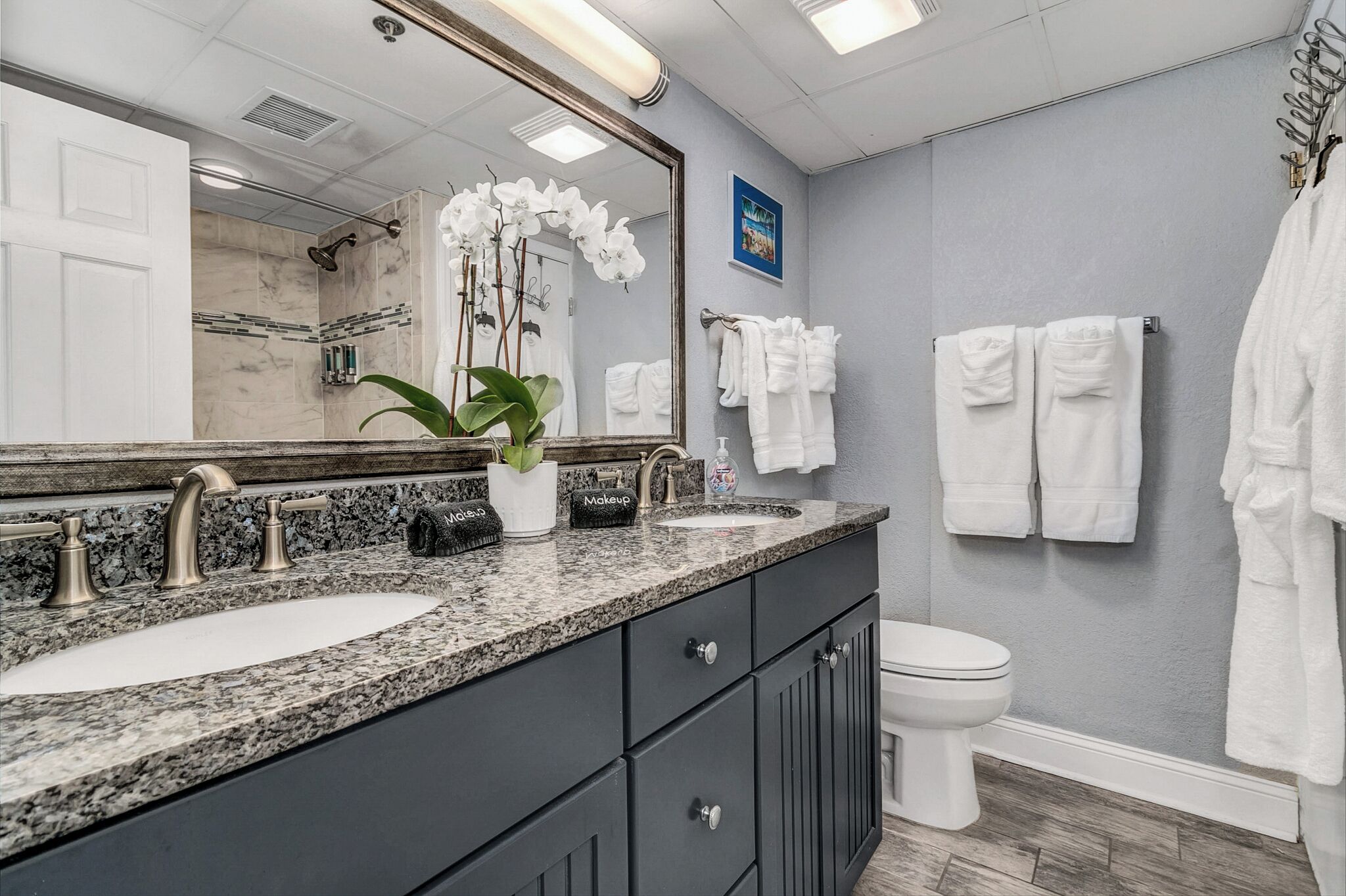 A bathroom with two sinks, a large mirror, and a toilet. There are towels hanging on the wall and a plant on the counter.