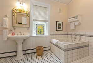 Casa de banho com lavatório pedestal, banheira, janela com persianas, quadro emoldurado na parede e porta-toalhas.