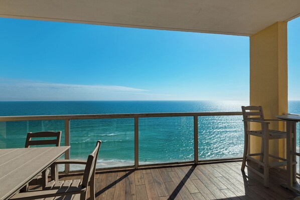 A balcony with a wooden floor, a table, and two chairs, overlooking a clear blue sea and a distant horizon.