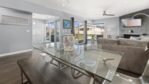 A modern living room with a glass-top dining table, a gray sofa, and a ceiling fan. - Stunning 6-Bedroom Oceanview Vacation Home w/ Rooftop Deck (Newport Beach)