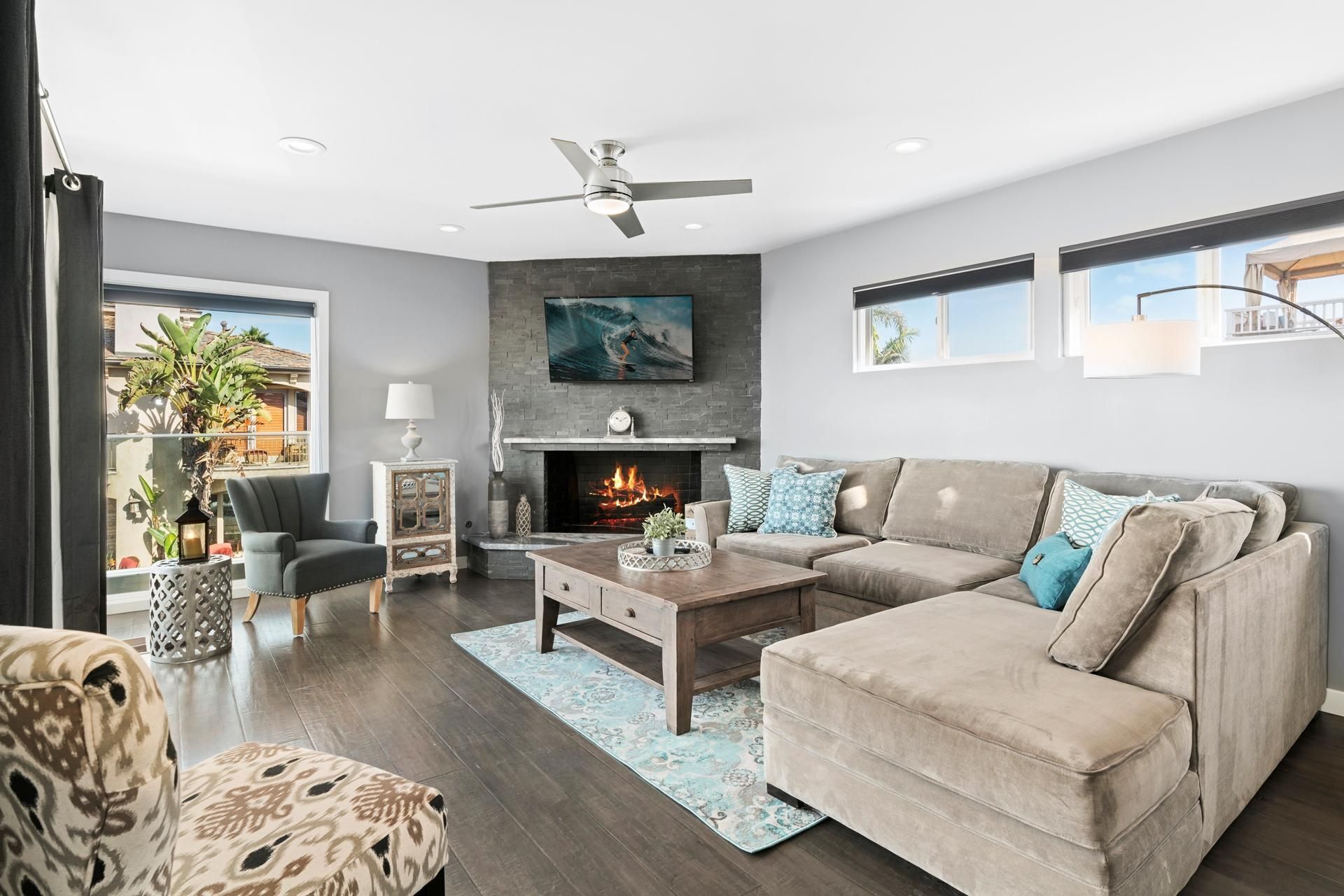 A modern living room with a fireplace, a ceiling fan, and a large window overlooking a garden.