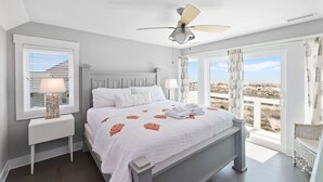 Ein Schlafzimmer mit Bett, Deckenventilator, Nachttisch, Lampe und Blick auf den Strand.