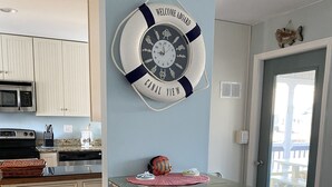 Una cucina con mobili bianchi, un tavolo con piano in vetro e due sgabelli da bar bianchi. Sulla parete è appeso un orologio a forma di salvagente e sulla porta c'è una decorazione a tema pesce.