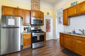A kitchen with wooden cabinets, stainless steel appliances, a brick wall, and a colorful painting.