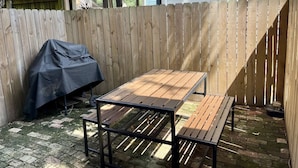 Ein Holztisch mit Bank in einem Hinterhof, umgeben von einem Holzzaun und einem überdachten Grill.