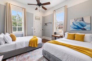 A hotel room with two beds, a ceiling fan, a painting, and a view of the outdoors.