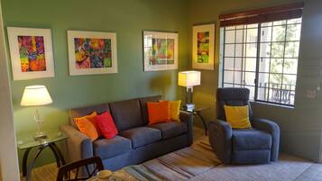 A living room with a blue sofa, two armchairs, a glass coffee table, and a window with a view of trees.