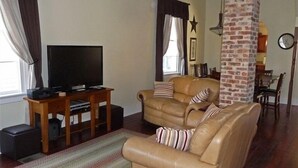 Un salon avec un canapé en cuir, un téléviseur posé sur un meuble en bois et un pilier en briques.