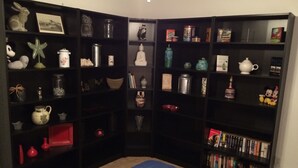 A room with a blue carpet, a bookshelf filled with books and decorative items, and a small white trash bin.