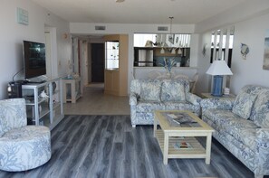 A living room with a sofa, armchair, coffee table, TV, and a kitchen area in the background.