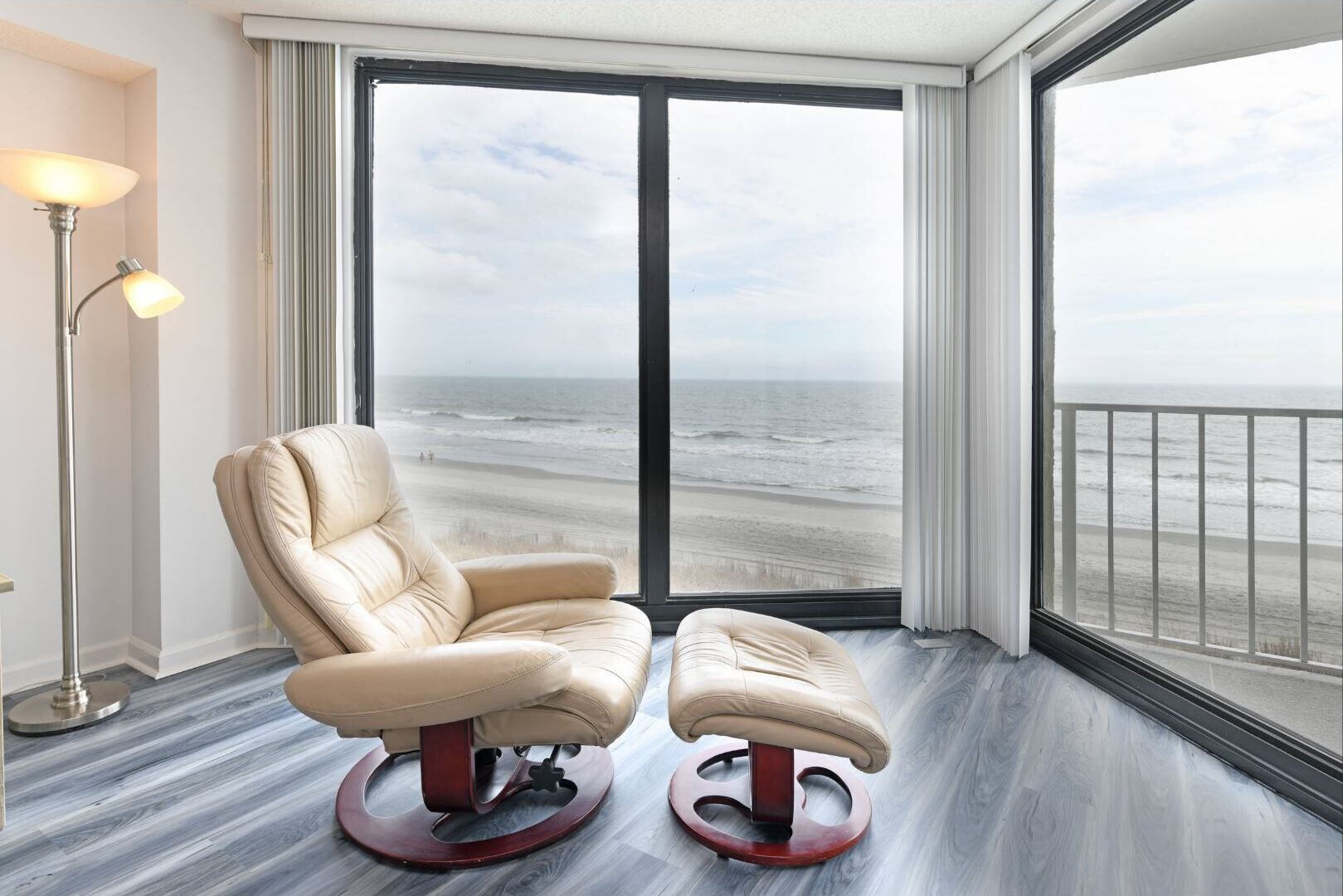 A room with a large window overlooking a beach, a beige leather chair, a small ottoman, and a floor lamp.