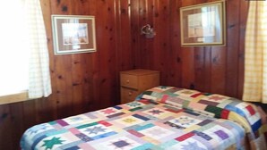 Ein kleiner, gemütlicher Raum mit Holzwänden, einem Einzelbett mit bunter Steppdecke, einem Nachttisch aus Holz und zwei gerahmten Bildern an der Wand.