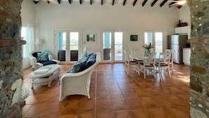 A spacious living area with a high ceiling, stone walls, and a tiled floor. There are two white wicker sofas with blue cushions, a dining table with chairs, and a kitchen area with a refrigerator and cabinets.