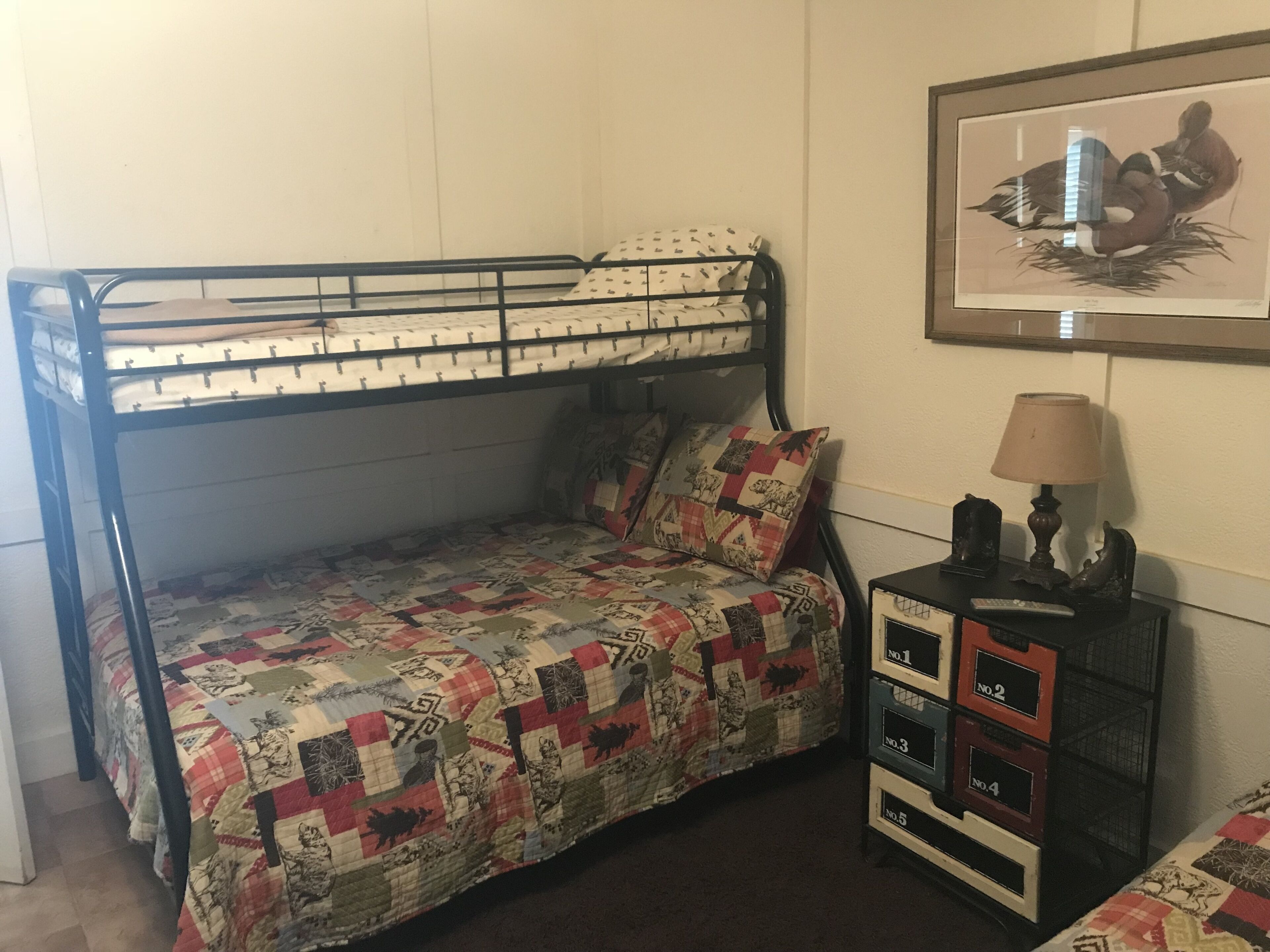 A bunk bed with a quilted bedspread, a bedside table with drawers, and a framed picture on the wall.
