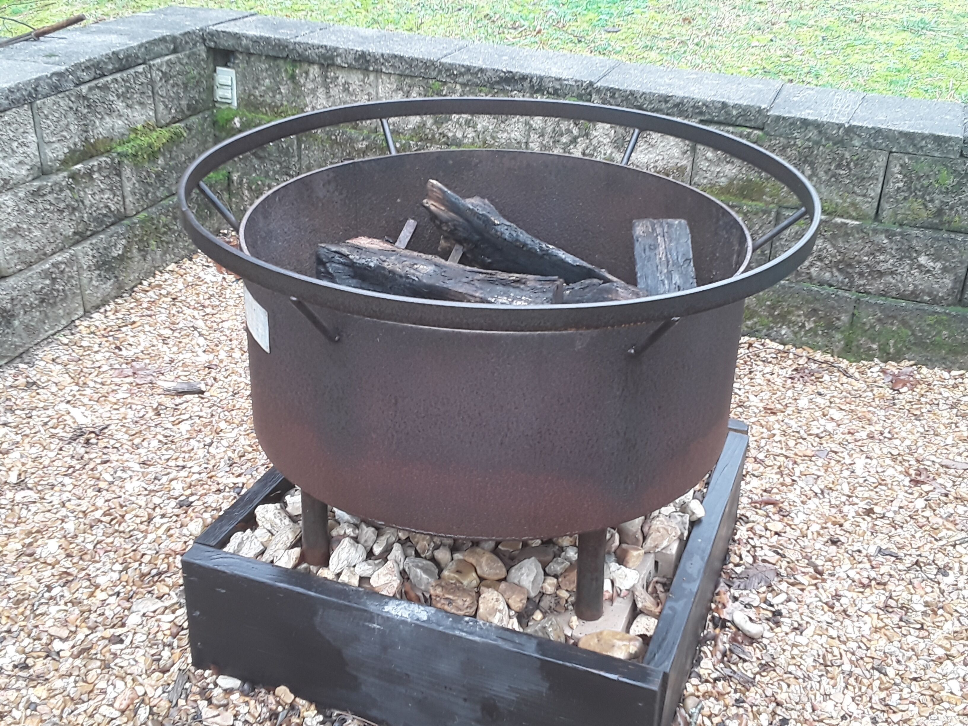 A metal fire pit with logs, surrounded by gravel, against a stone wall.