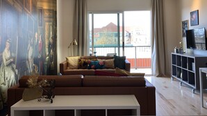 Un salon avec un grand tableau, un canapé marron, une table basse blanche et une vue sur les immeubles à travers de grandes fenêtres.
