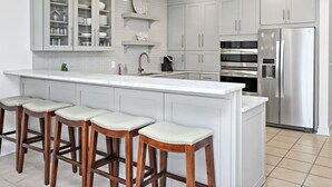 A modern kitchen with white cabinetry, a stainless steel refrigerator, and a bar area with stools. - Gorgeous Overlooking Gulf,Patio, Pool, Grill (Santa Rosa Beach)