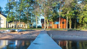 Een betonnen pad dat leidt naar een houten hutje aan een waterpartij, omringd door bomen en boten.