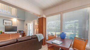 Un salon chaleureux avec un canapé en cuir marron, une table à manger en bois, des chaises en osier et une grande fenêtre équipée de stores.