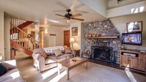 A spacious living room with a stone fireplace, a ceiling fan, a television, and a staircase leading to another level. - (12) Cabin Twelve at The Redwoods In Yosemite (Wawona)