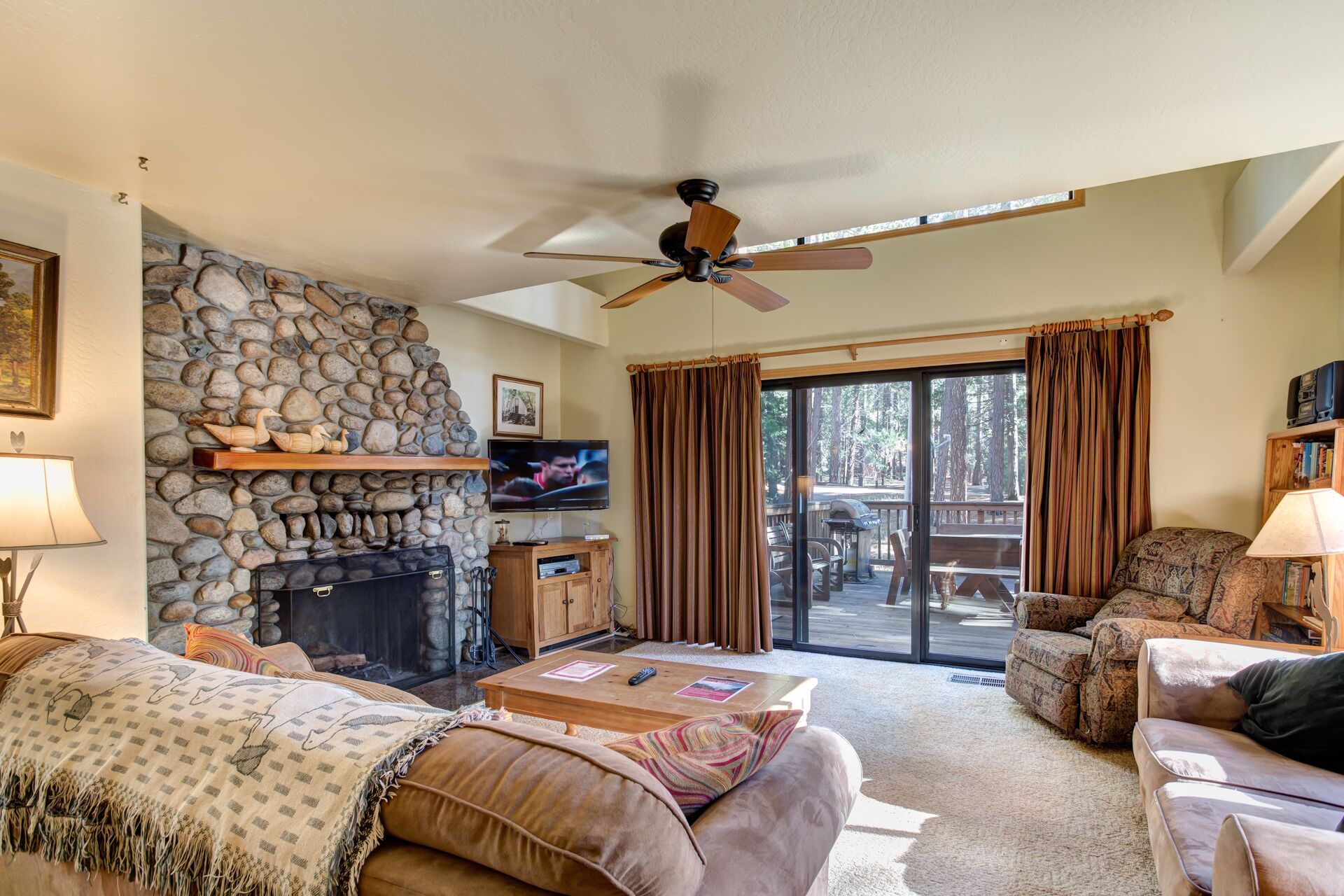 Una sala de estar acogedora con chimenea de piedra, ventilador de techo y vistas al exterior.