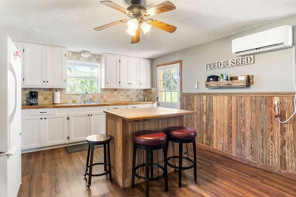 A kitchen with white cabinets, a wooden island with stools, a ceiling fan, and an air conditioning unit.