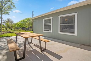A patio with a wooden table and benches, a grey building with two windows, and a green lawn.