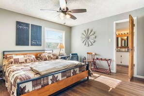 A bedroom with a bed, a ceiling fan, a window, a wall clock, and a door leading to a bathroom.