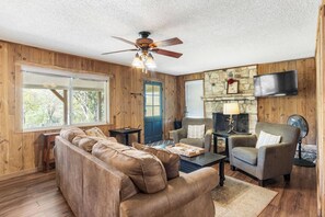 A cozy living room with a sofa, armchairs, a coffee table, a fireplace, a television, and a ceiling fan.