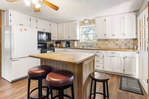A kitchen with white cabinets, a wooden island, two stools, a refrigerator, a microwave, and a window with a view.