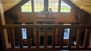 L’intérieur d’une cabane en rondins, avec un plafond haut, une cheminée en pierre et une grande fenêtre donnant sur un paysage verdoyant.